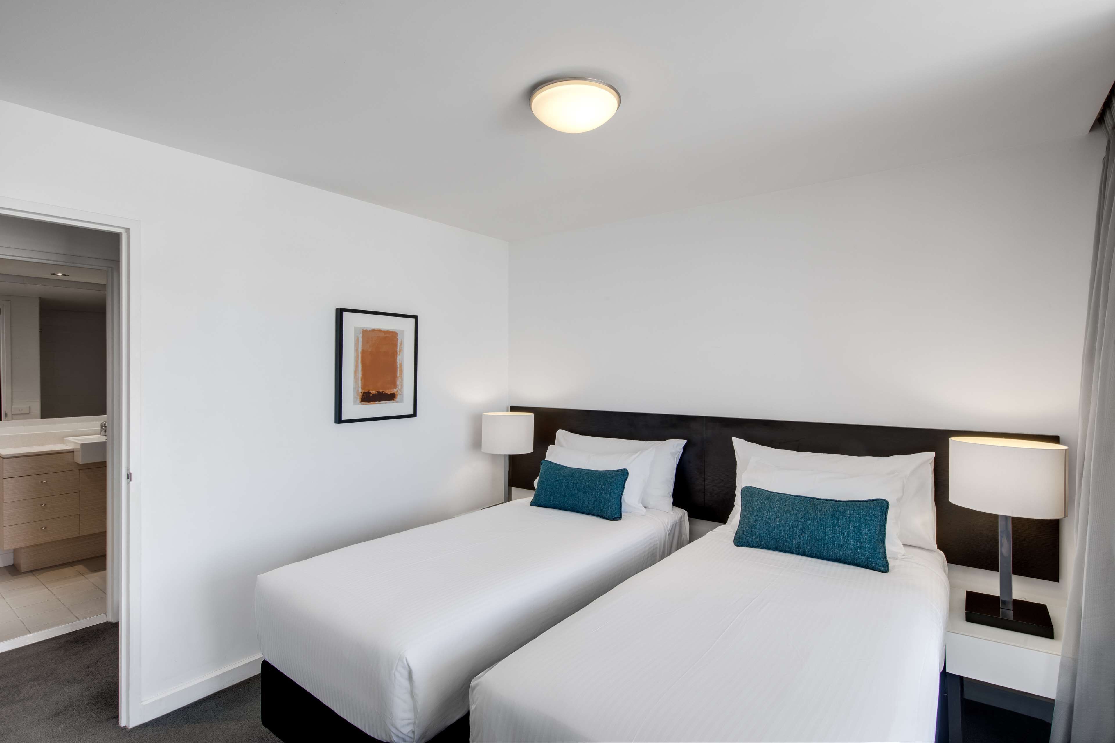 A hotel room with two single beds, a bedside table with a lamp, a framed picture on the wall, and a bathroom visible through an open door.