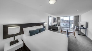 Moderni hotellihuone, jossa on suuri sänky, työpöytä ja televisio, oleskelualue pöydällä ja tuoleilla sekä parveke, jolta on näkymä kaupunkiin.