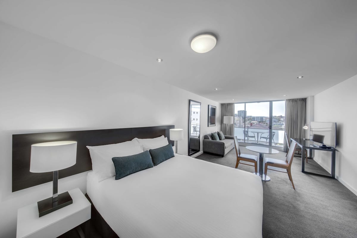 Ett modernt hotellrum med en stor säng, ett skrivbord med tv, en sittgrupp med bord och stolar samt en balkong med utsikt över staden.