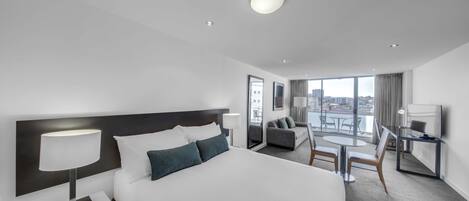 A modern hotel room with a large bed, a desk with a TV, a seating area with a table and chairs, and a balcony with a city view.