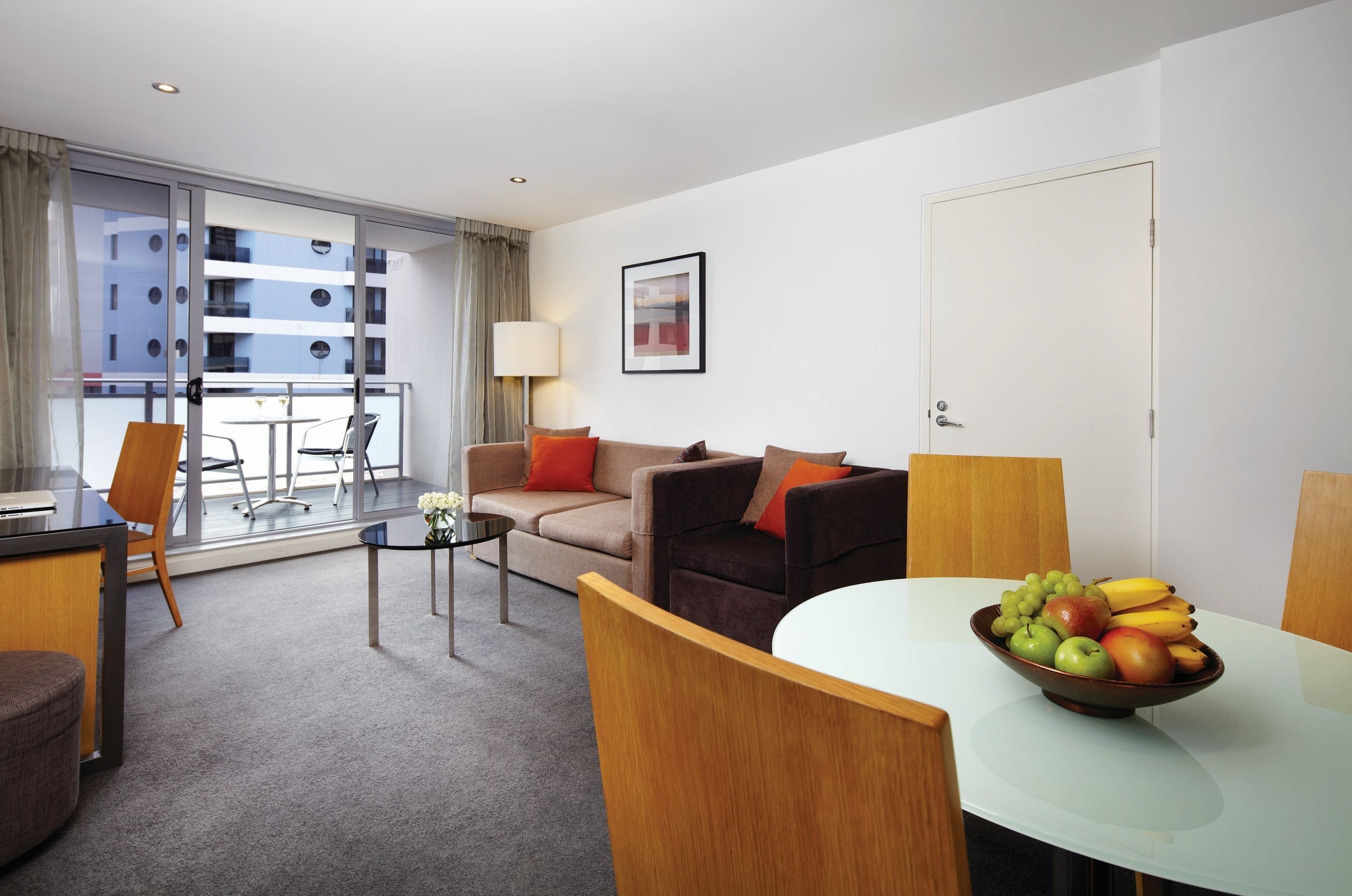 Quarto de hotel com sofá, mesa de jantar com tigela de frutas, escrivaninha e cadeira. Possui uma varanda com vista para edifícios.
