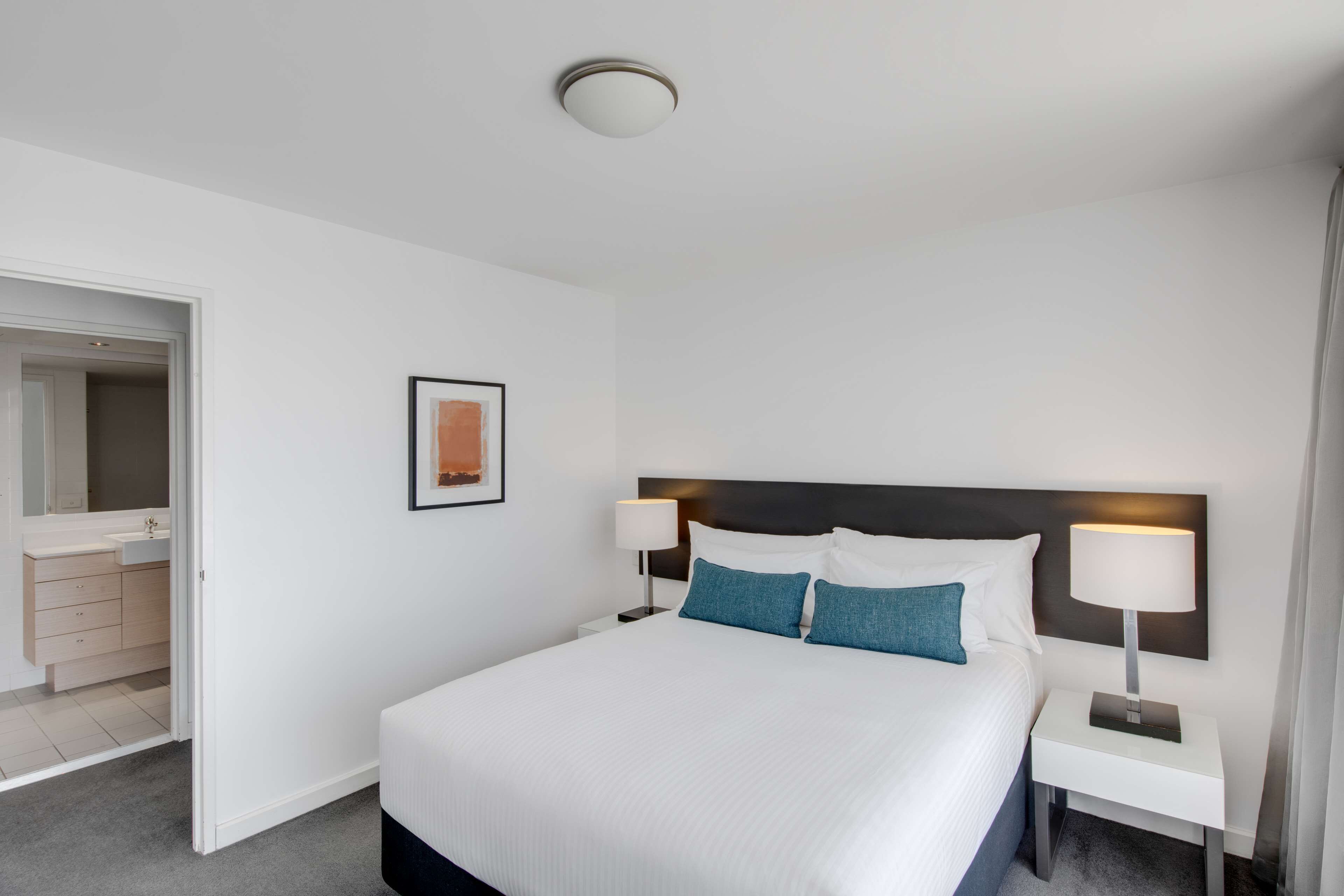 Hotelkamer met een groot bed, witte muren, een inlijsting, een nachtkastje met een lamp en een badkamer zichtbaar door een open deur.