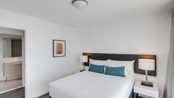 A hotel room with a large bed, white walls, a framed picture, a nightstand with a lamp, and a bathroom visible through an open door.