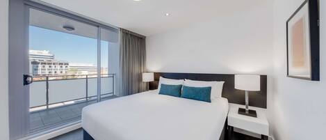 Habitación de hotel con una cama grande, una mesita de noche con lámpara, vista a la ciudad desde un balcón y una luz en el techo.