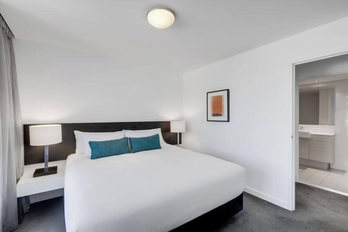 Ett hotellrum med en stor säng, ett nattduksbord med en lampa, en inramad tavla på väggen och en öppen dörr som leder till ett badrum.