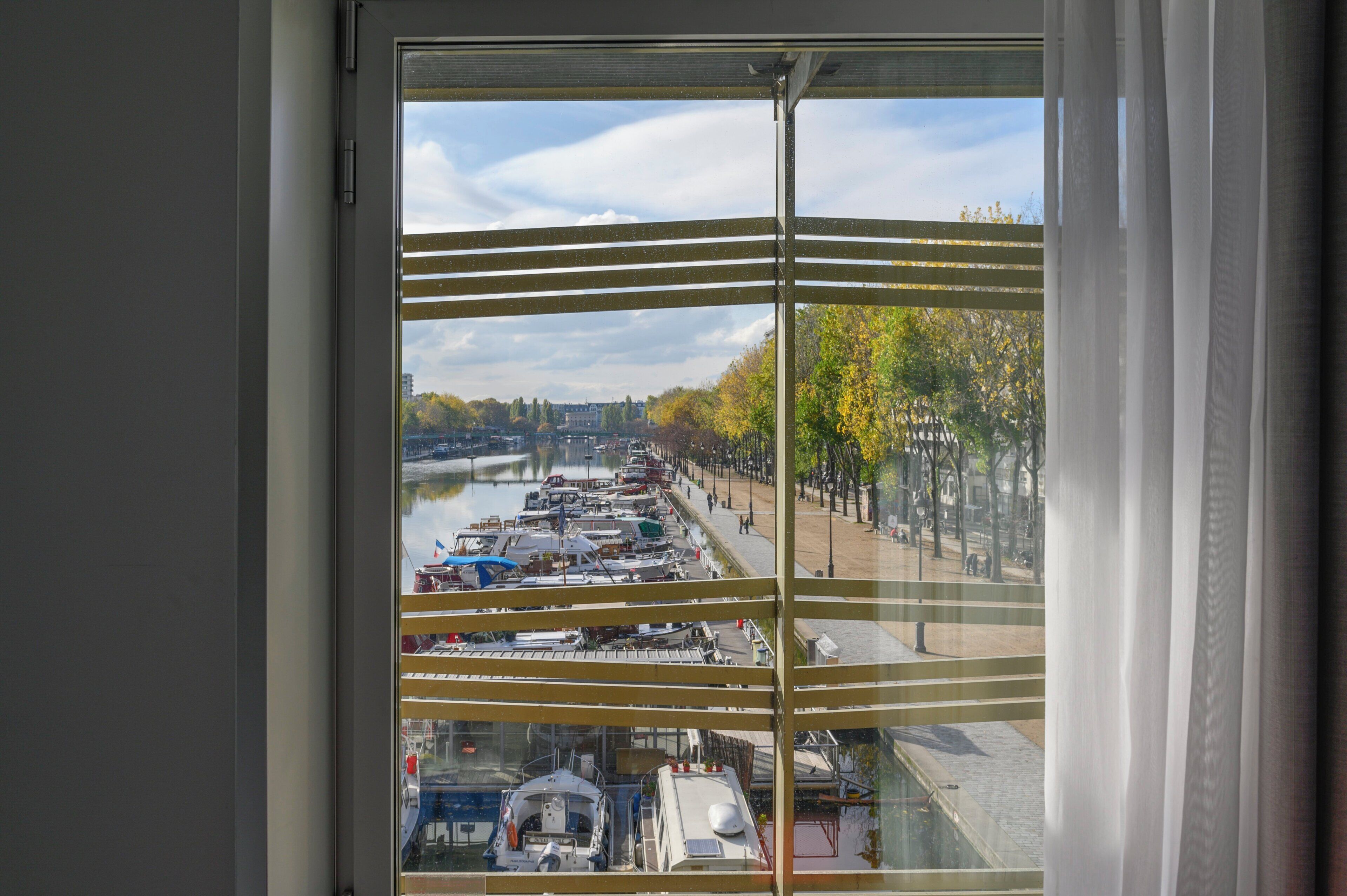 Uitzicht door een raam met horizontale zonwering, op een rivier met aangemeerde boten en een promenade met bomen.