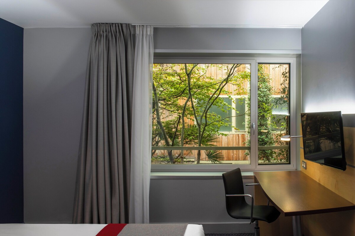 A hotel room with a bed, a desk, a chair, and a window with a view of trees and a fence.