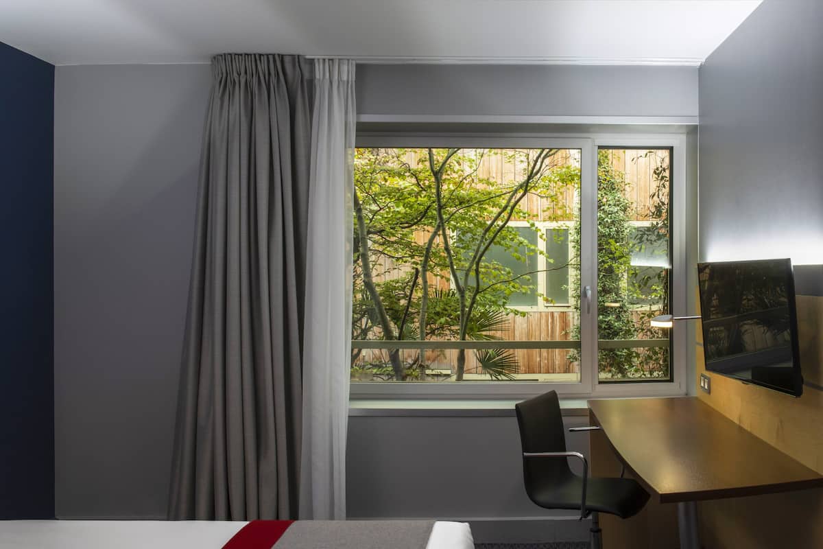 A hotel room with a bed, a desk, a chair, and a window with a view of trees and a fence.