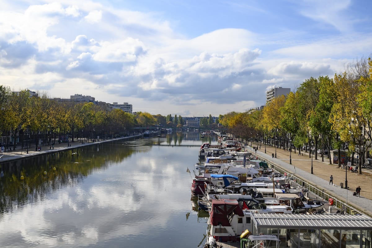 A riverside view with boats docked along the riverbank, a bridge in the distance, and trees lining the path.