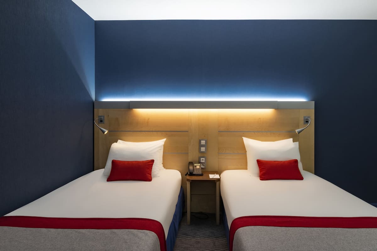 A hotel room with two beds, each with white bedding and red pillows, a wooden headboard, a bedside table with a lamp, and a wall-mounted light fixture.