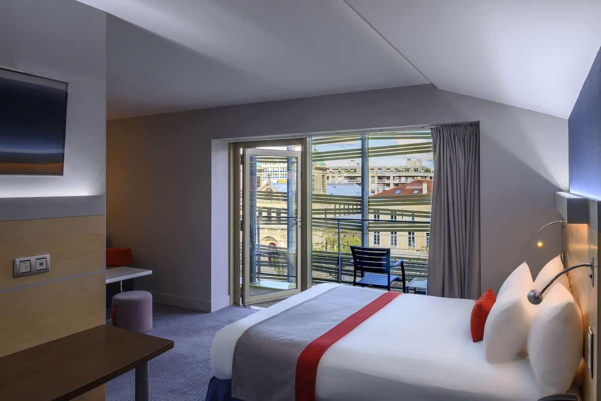 A hotel room with a bed, a desk, a chair, a TV, and a balcony with a view of buildings.