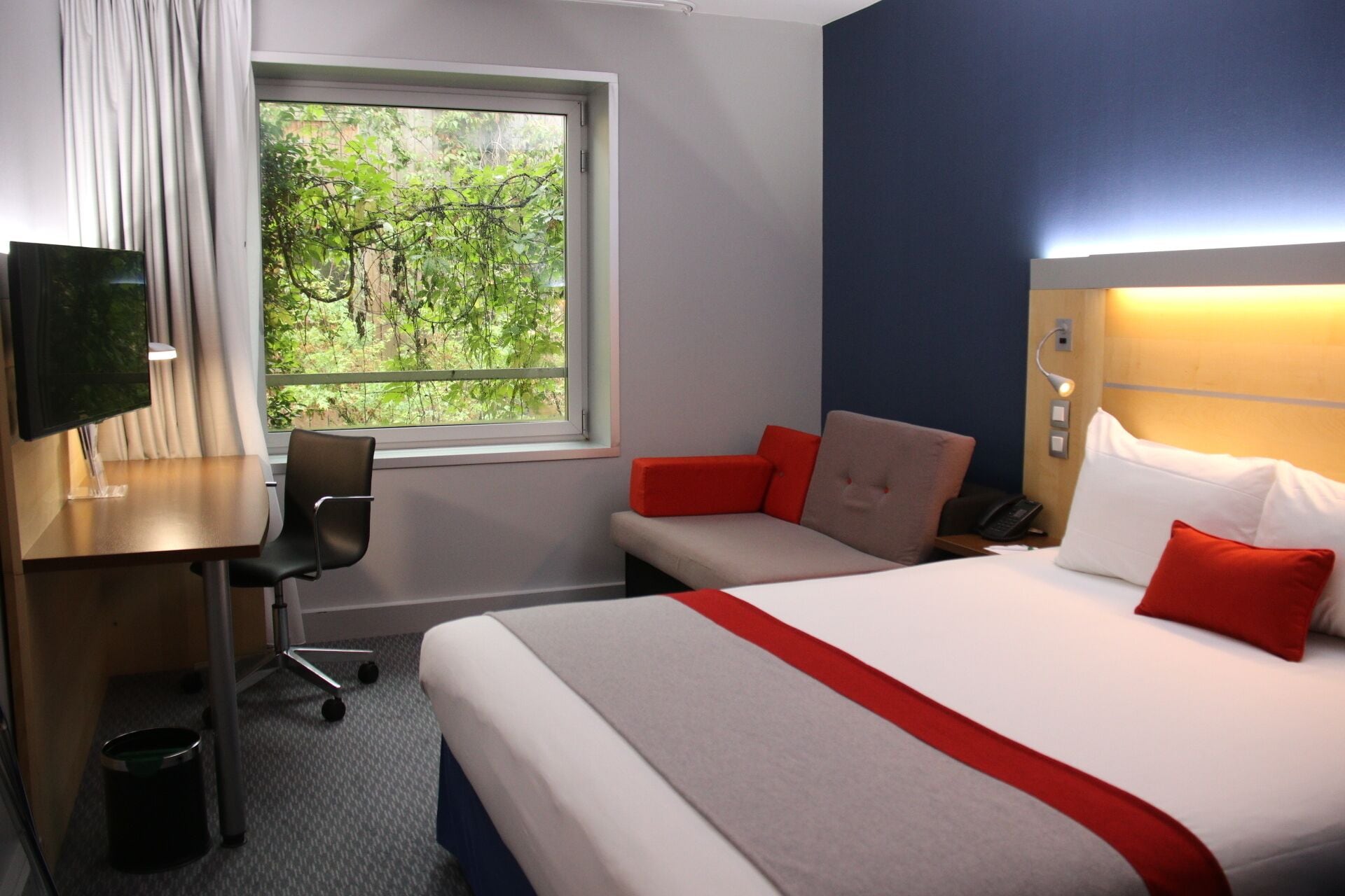 Habitación de hotel con cama, escritorio, silla, televisor y ventana con vistas a zonas verdes.