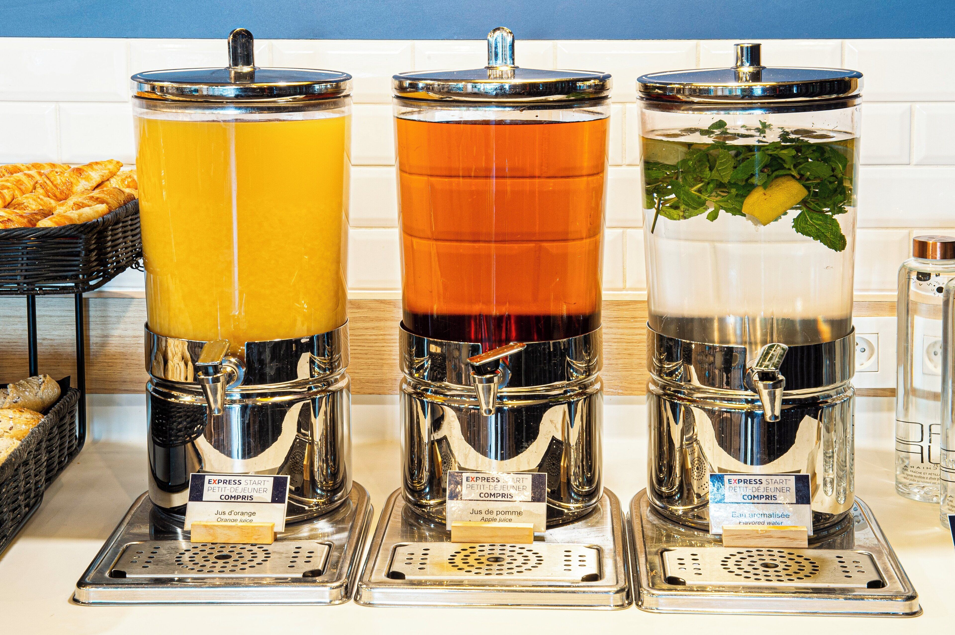 Trois distributeurs de boissons, chacun portant une étiquette indiquant le type de boisson : jus d'orange, jus de pomme et eau aromatisée.