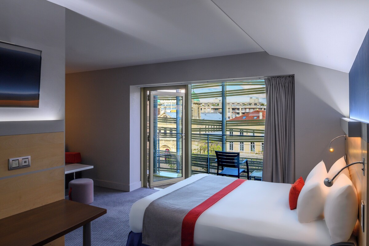 A hotel room with a bed, a desk, a chair, a TV, and a balcony with a view of buildings.