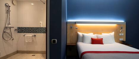 Quarto de hotel com chuveiro, cama com roupa de cama branca e um travesseiro vermelho, e mesa de cabeceira com abajur.