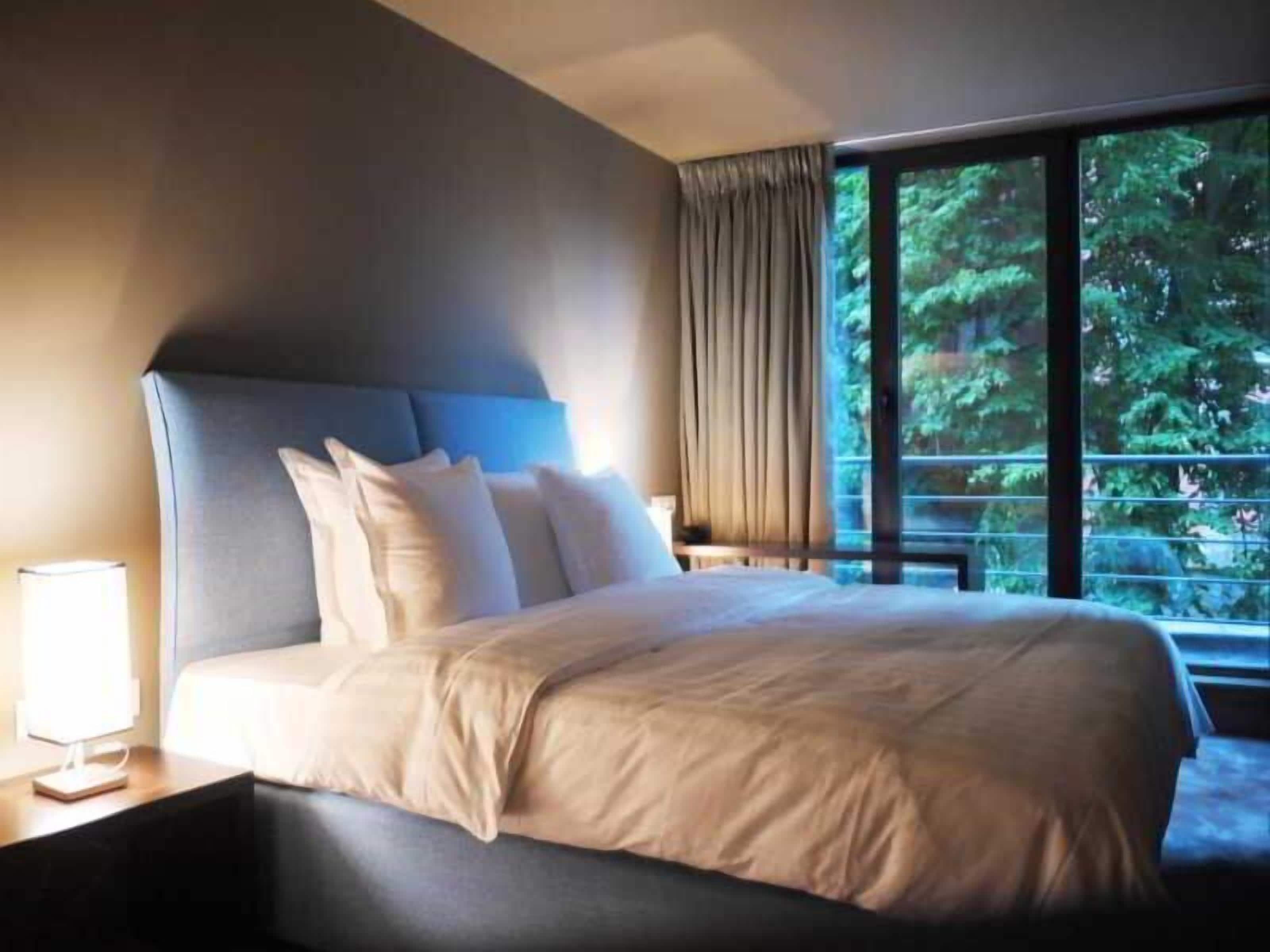 A hotel room with a large bed, a nightstand, a lamp, and a window with a view of trees.