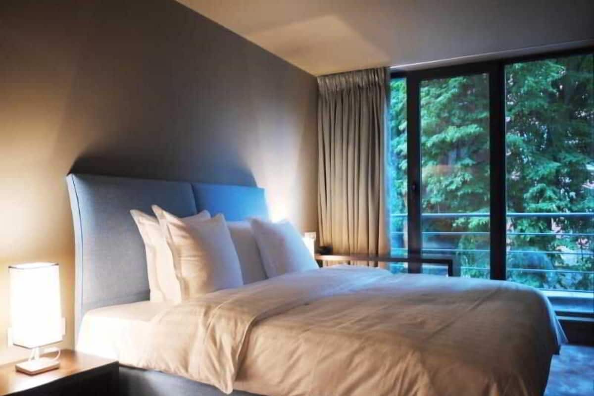 A hotel room with a large bed, a nightstand, a lamp, and a window with a view of trees.