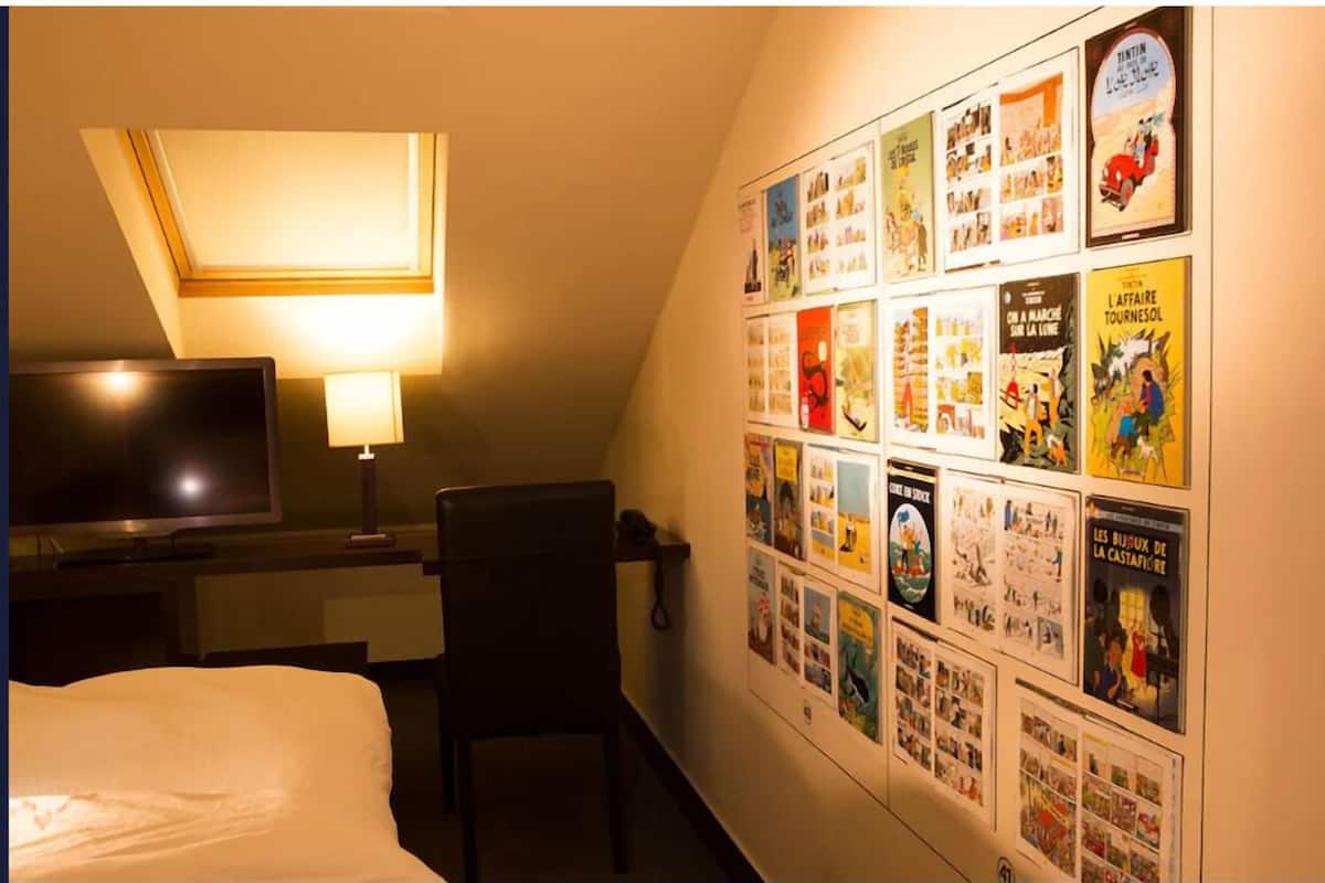 A hotel room with a bed, a desk with a lamp, a TV, and a wall decorated with various posters.