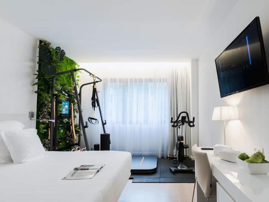 Una habitación de hotel moderna con una cama, equipo de ejercicio, televisor y un escritorio con una lámpara y un frutero.