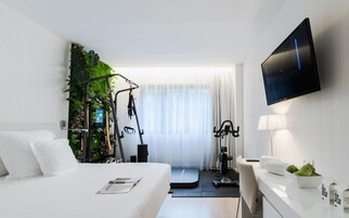 Camera d'albergo moderna con un letto, attrezzature per l'esercizio fisico, una TV e una scrivania con una lampada e una ciotola di frutta.