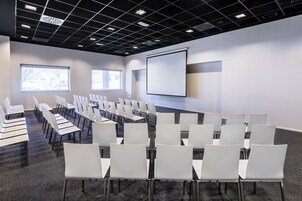 Ein Konferenzraum mit Reihen weißer Stühle, einer Projektionsleinwand und großen Fenstern.