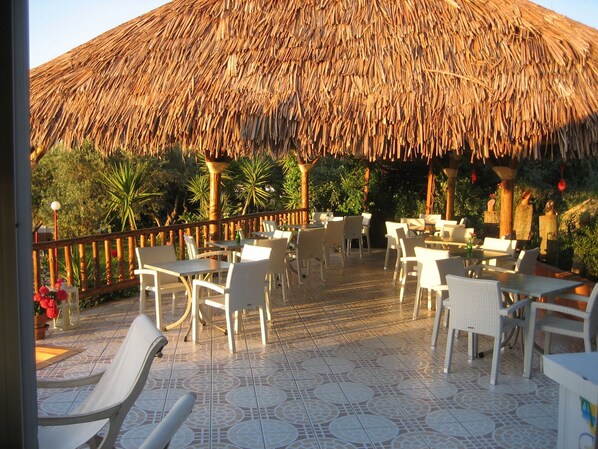 An outdoor patio with thatched roof seating and tables, surrounded by greenery. - New Kydonia Suites & Studios (Chania)