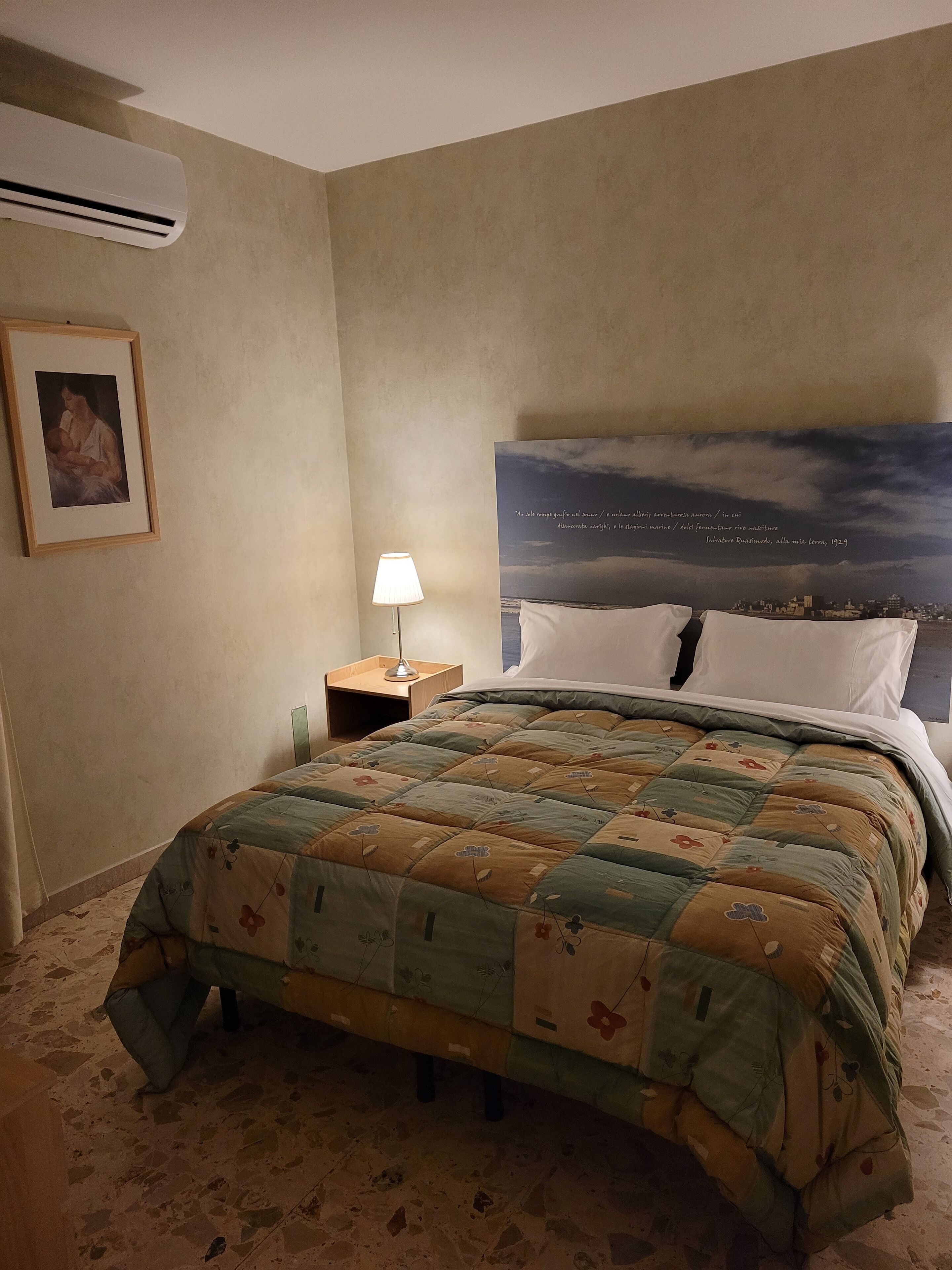 A hotel room with a bed, a nightstand with a lamp, a framed picture on the wall, and a large poster above the bed.