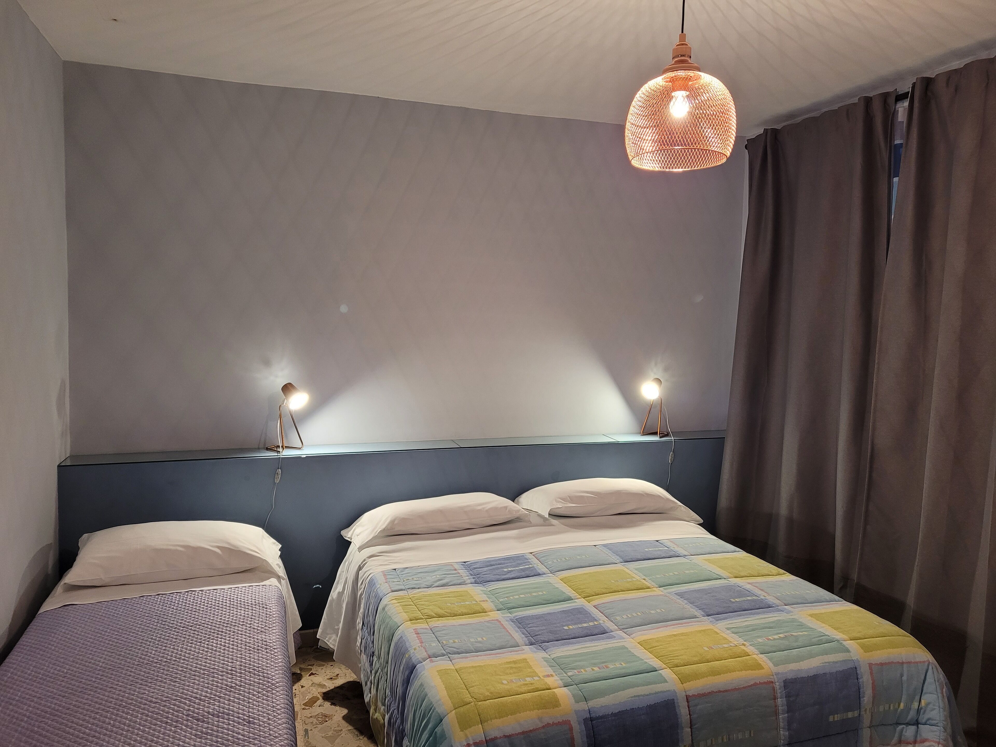 Une chambre avec deux lits simples, une tête de lit bleue, une couvre-lit matelassée aux couleurs vives et un luminaire suspendu.