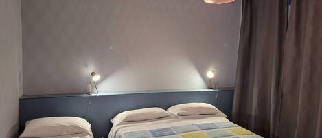 Une chambre avec deux lits simples, une tête de lit bleue, une couvre-lit matelassée aux couleurs vives et un luminaire suspendu.