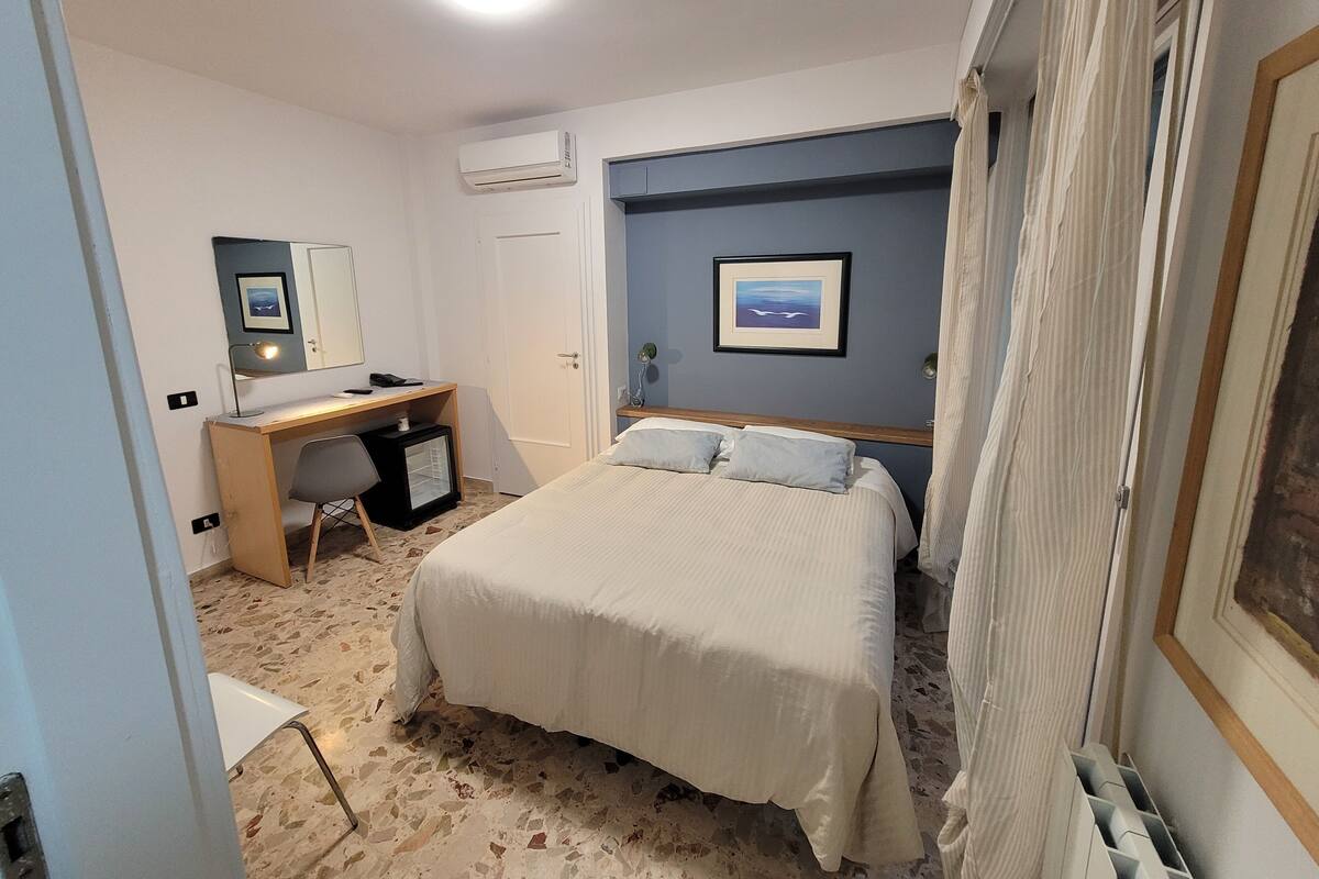 A bedroom with a bed, a desk, a chair, an air conditioner, and a framed picture on the wall.