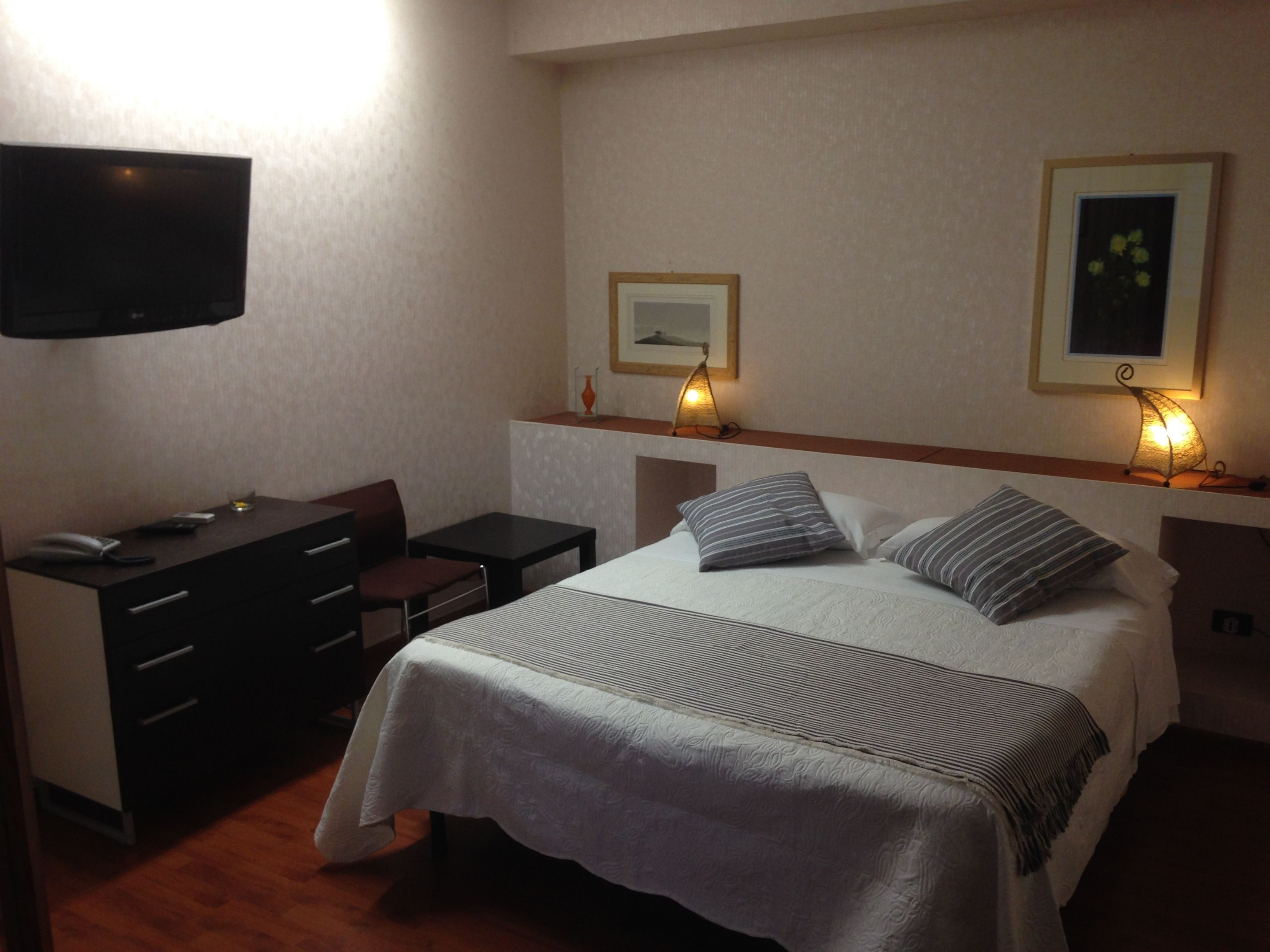 Habitación de hotel con una cama, un escritorio, una silla, un televisor y dos lámparas de pared.