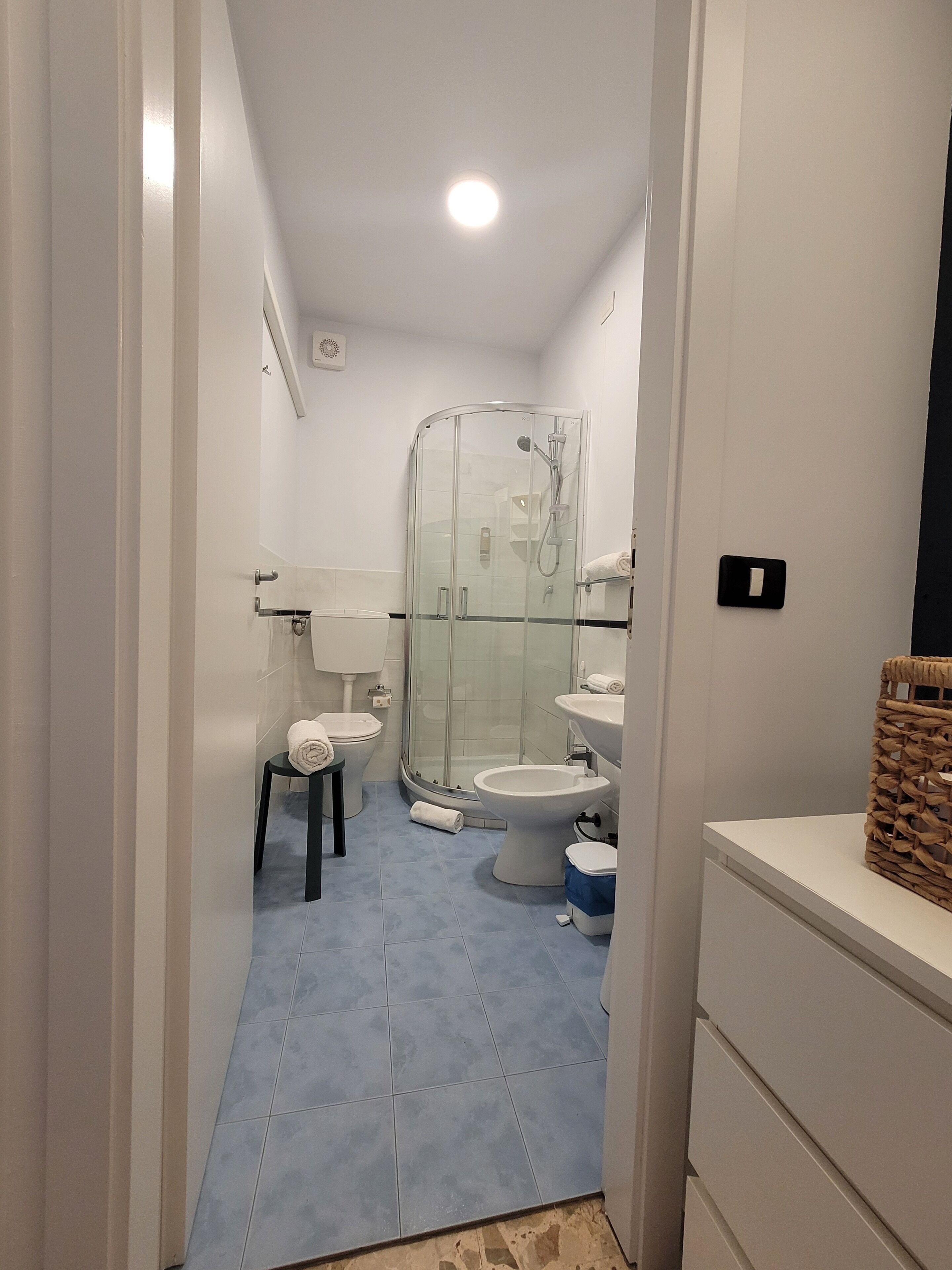 A bathroom with a glass shower enclosure, a toilet, a sink, and a small stool with a towel.