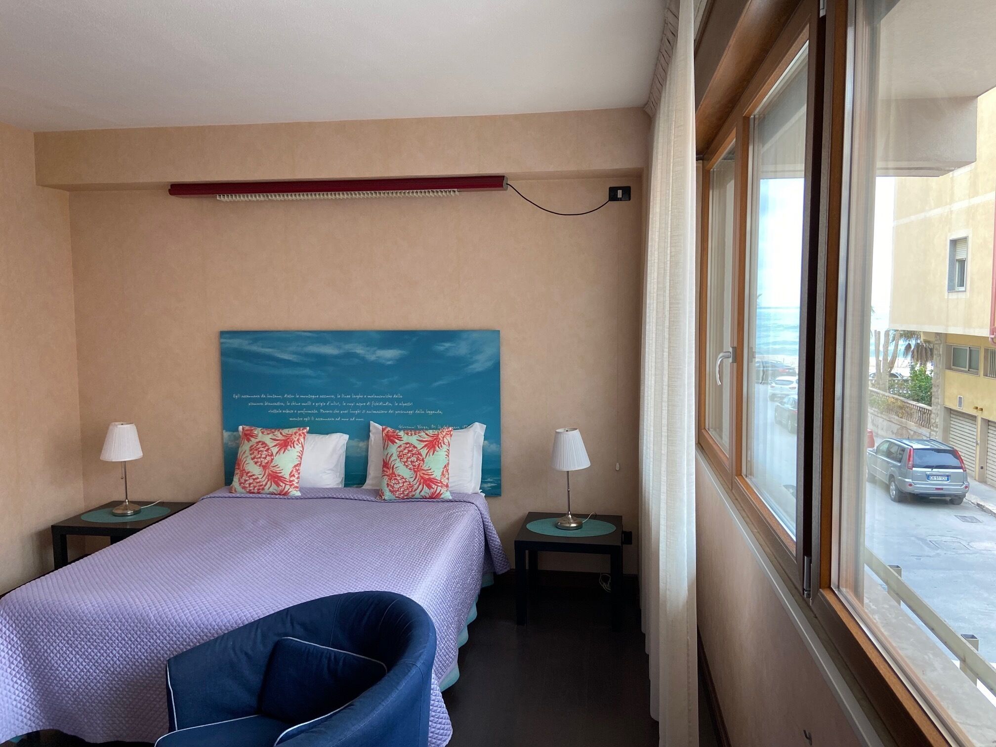 A hotel room with a bed, two bedside tables, a chair, a window with a view of the street, and a wall with a decorative poster.