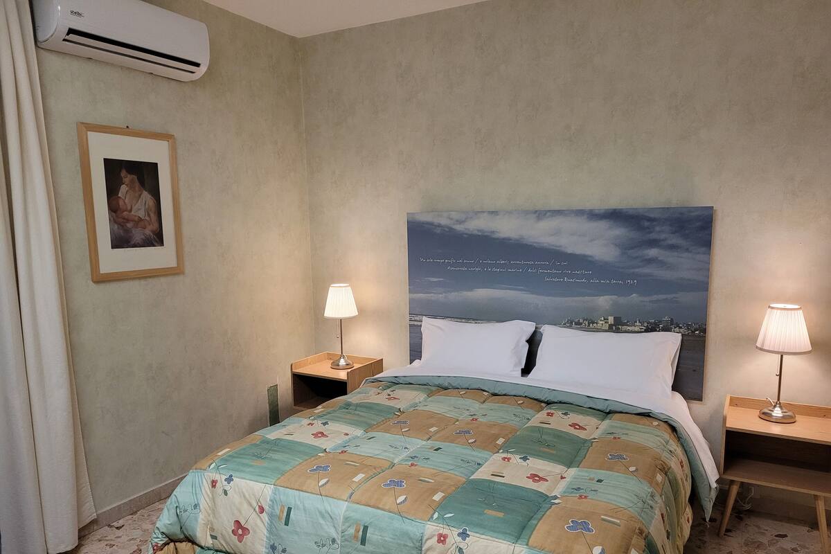 A hotel room with a bed, two bedside lamps, a framed picture on the wall, and a large window with a cityscape view.