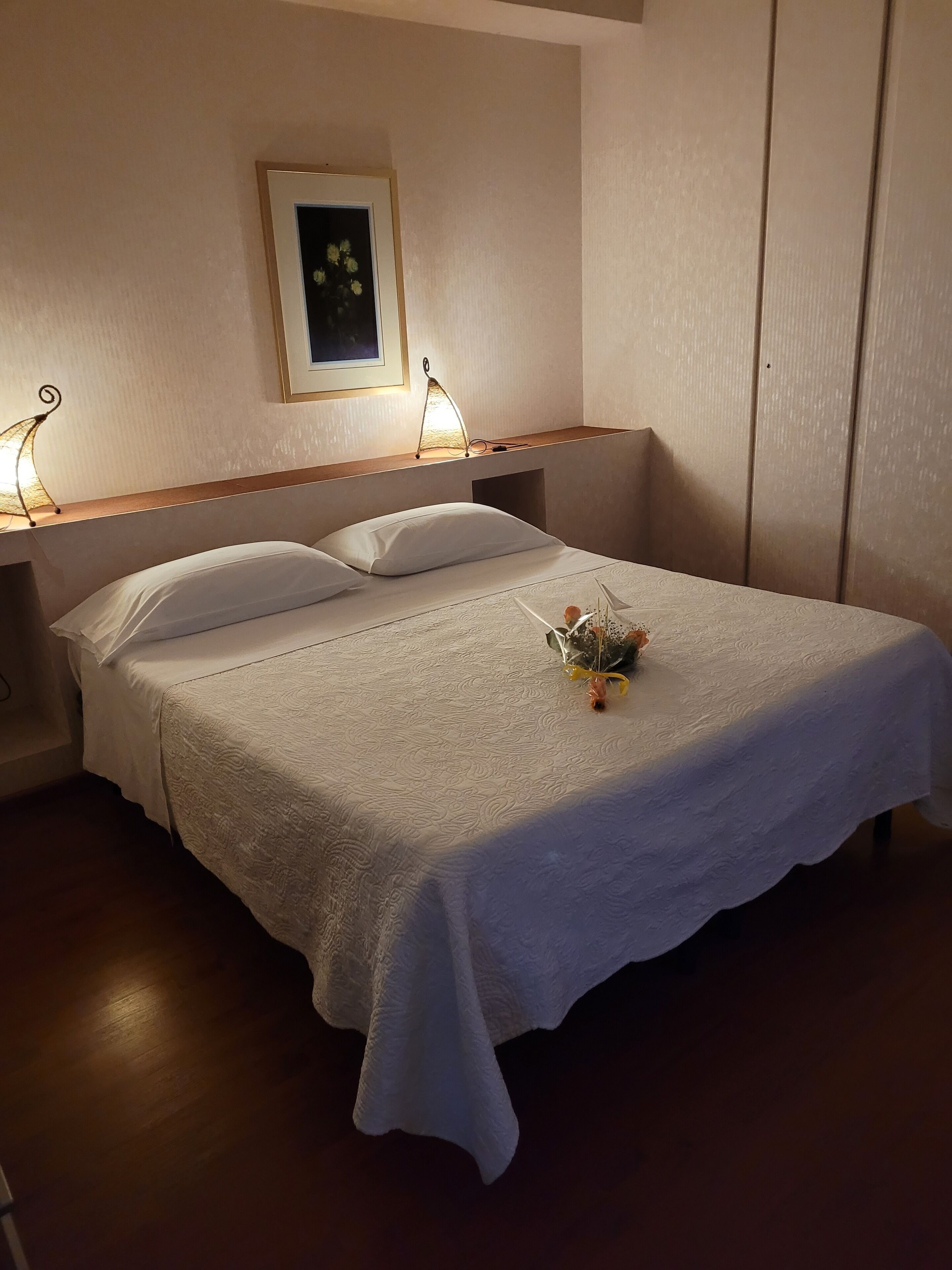 Una cama bien hecha con cubrecama y almohadas blancas, una mesita de noche con lámpara y un cuadro en la pared.