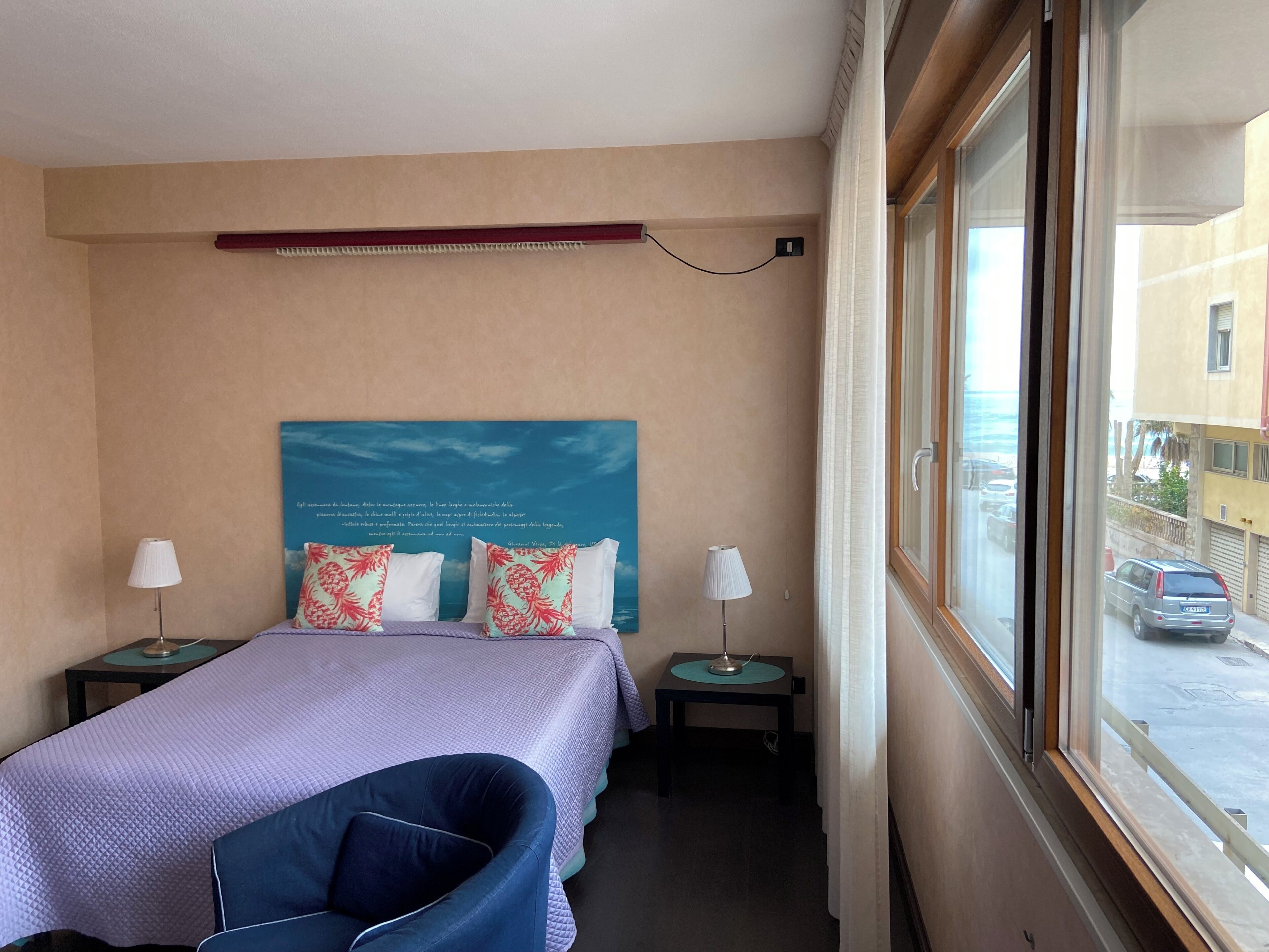 A hotel room with a bed, two bedside tables, a chair, a window with a view of the street, and a wall with a decorative piece.