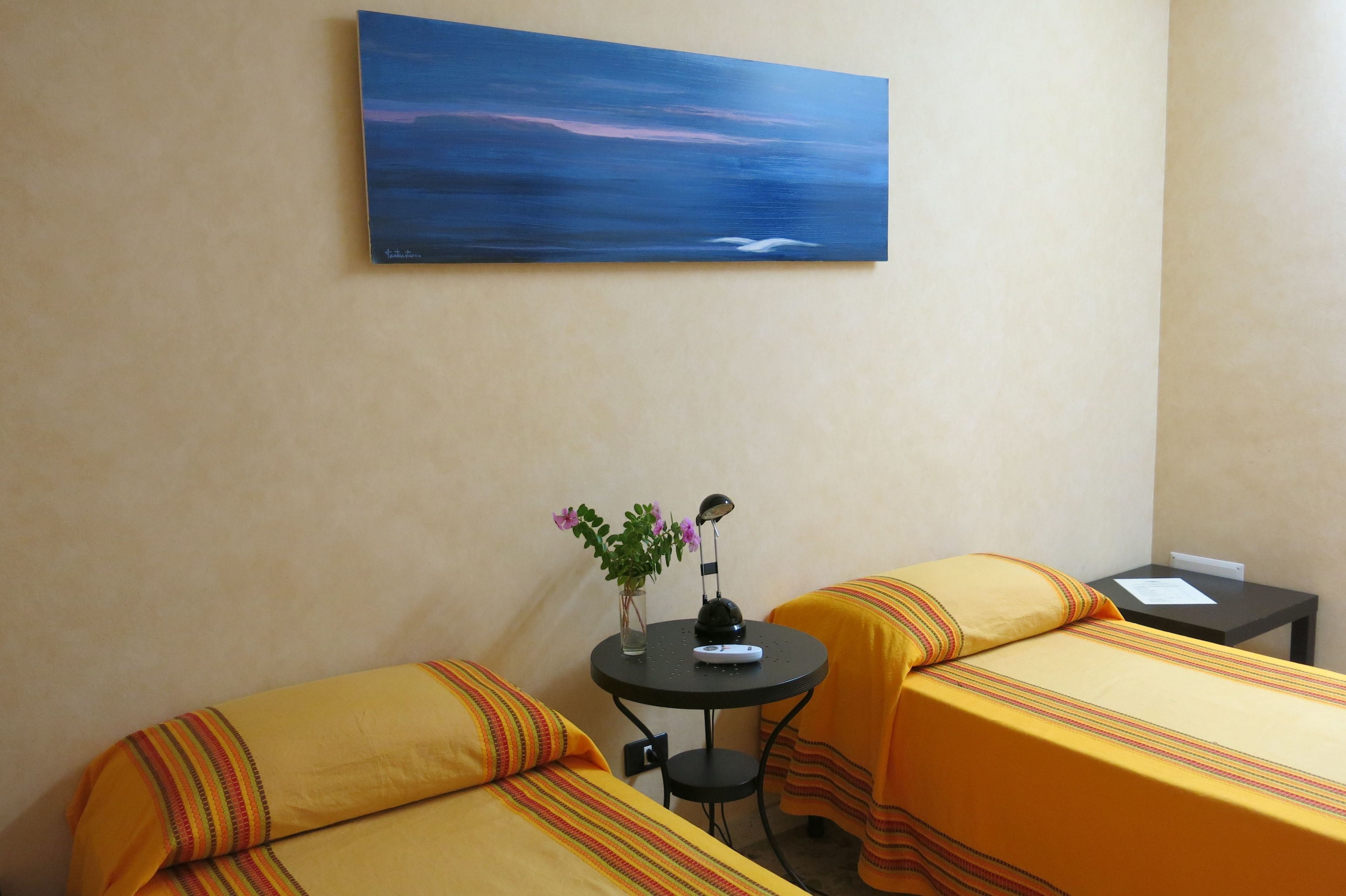 A hotel room with two beds, a small table with a lamp, a vase with flowers, and a painting on the wall.