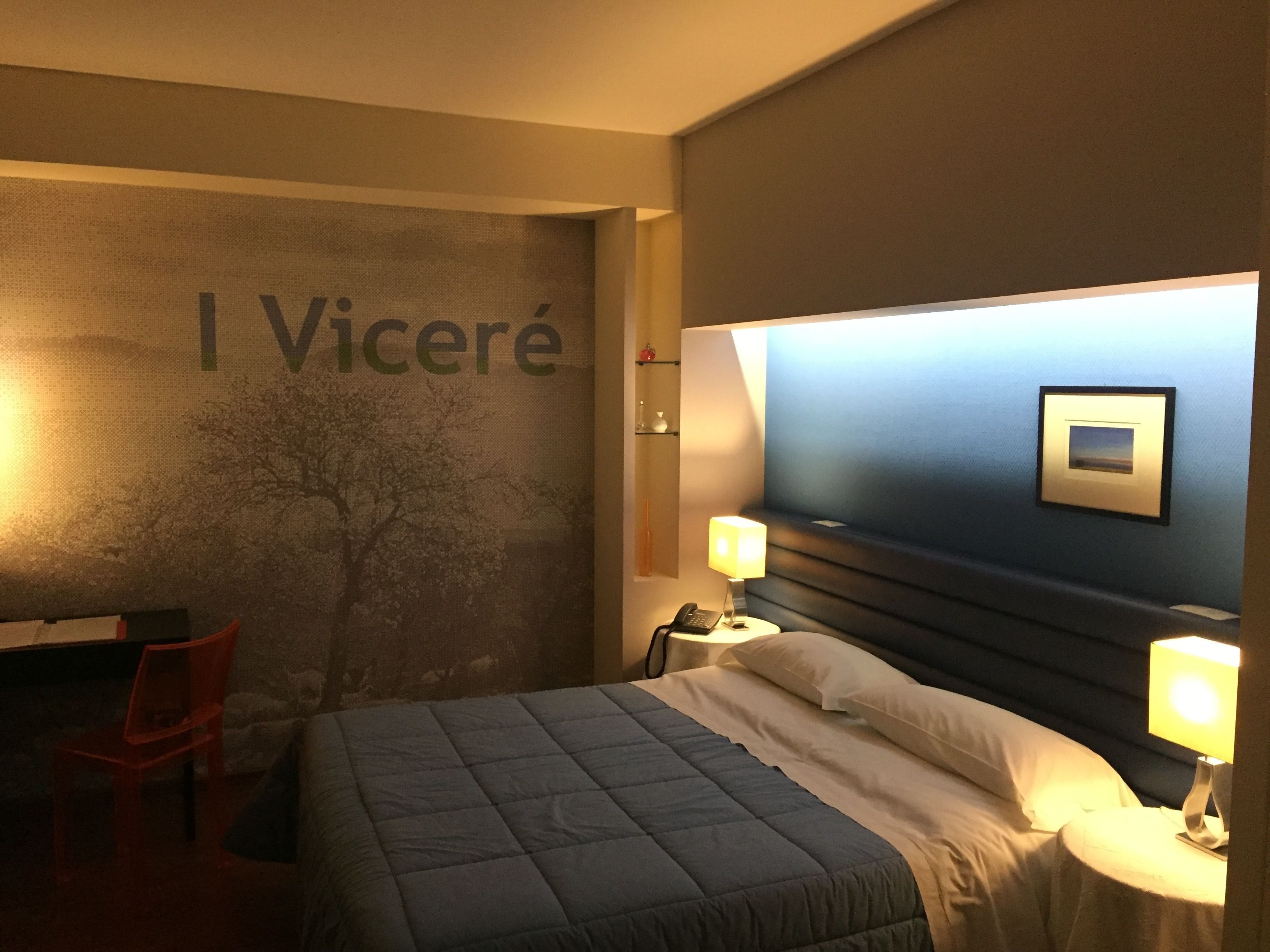 A hotel room with a bed, bedside lamps, a desk, and a chair. The wall has a large sign with the name 'I Viceré'.