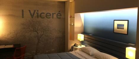 A hotel room with a bed, bedside lamps, a desk, and a chair. The wall has a large sign with the name 'I Viceré'.