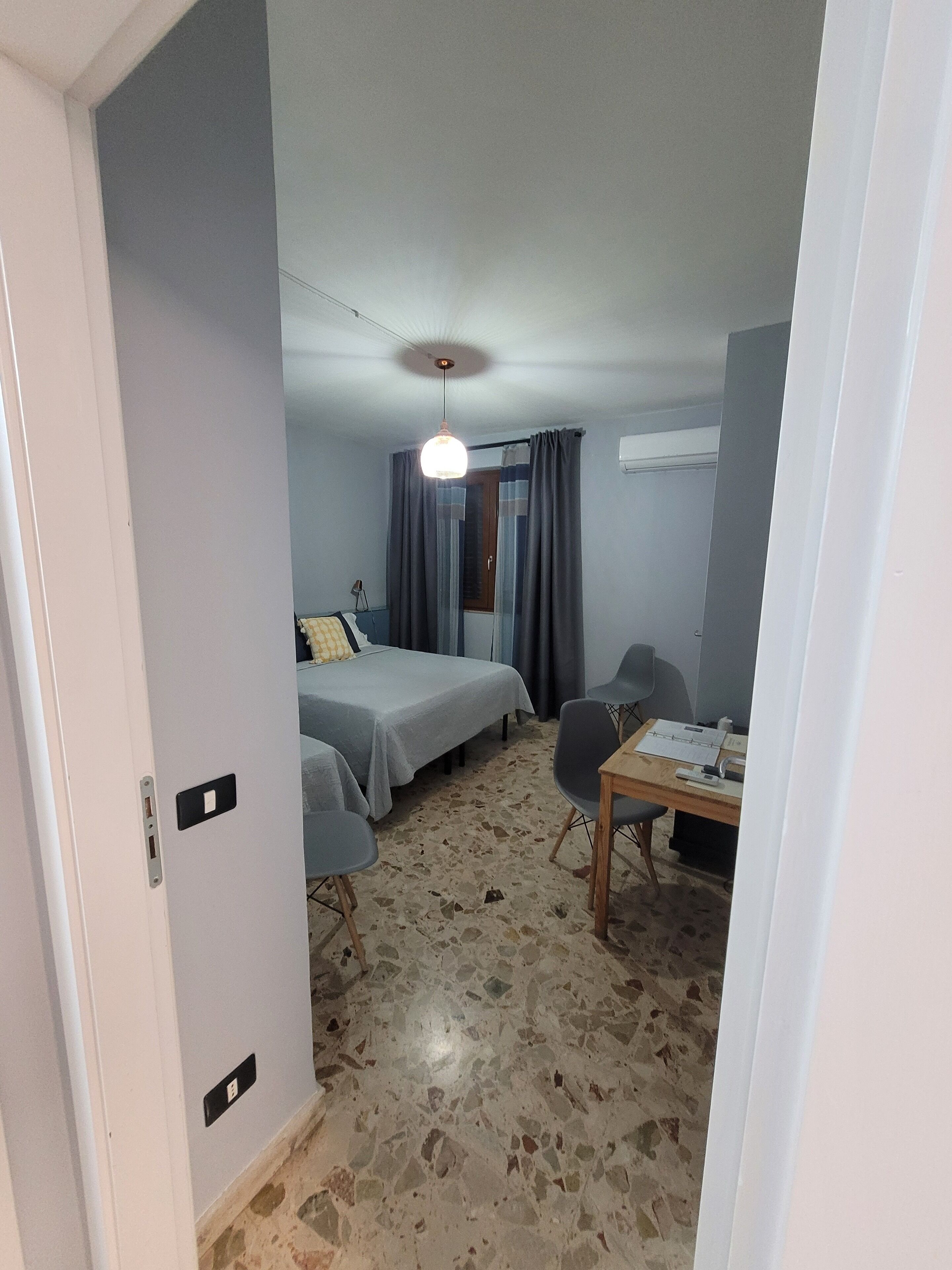 A room with two beds, a desk, and a chair. There is a ceiling light and a window with curtains.