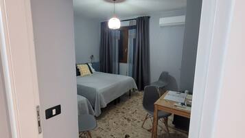 A room with two beds, a desk, and a chair. There is a ceiling light and a window with curtains.