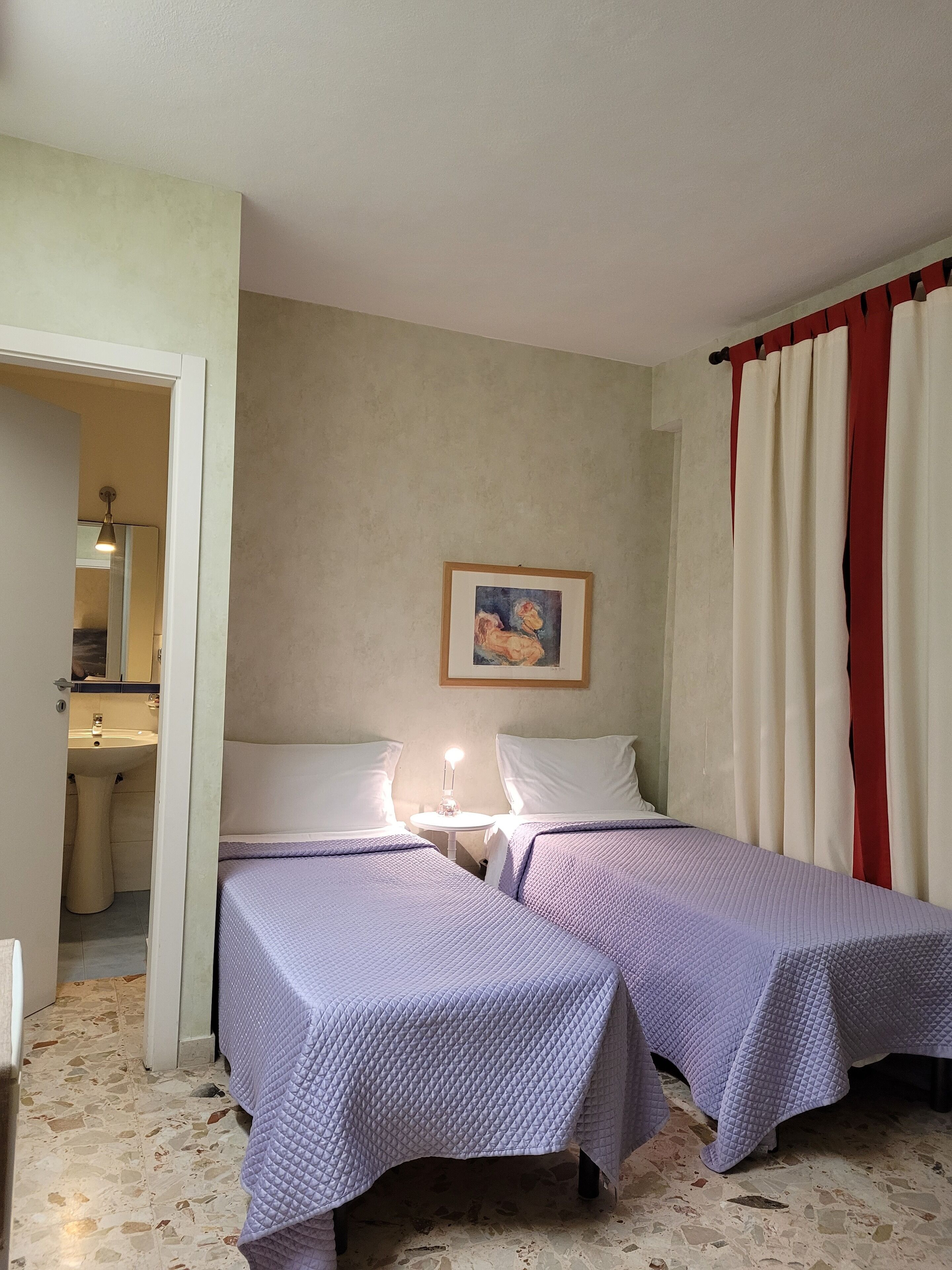A room with two beds, a painting on the wall, and a bathroom visible through an open door.