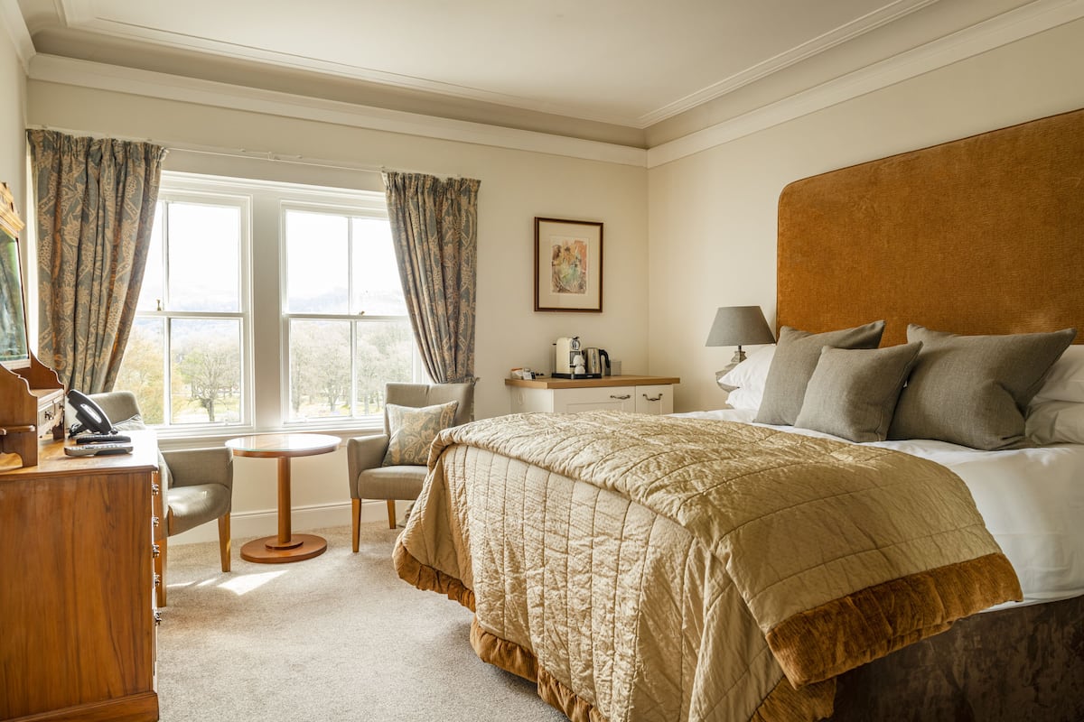 A hotel room with a large bed, a desk, a chair, a small table, and a window with curtains.