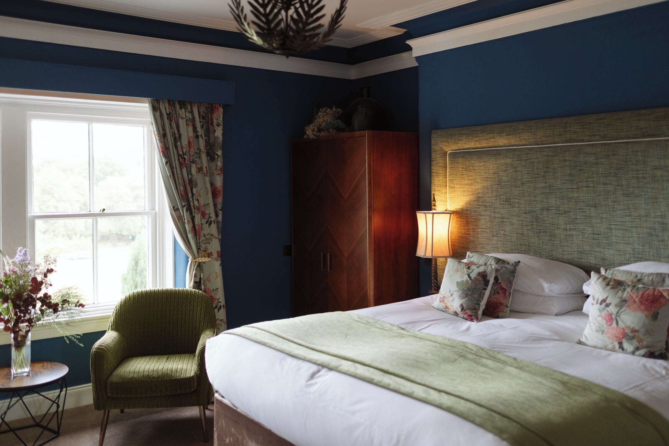 Een slaapkamer met een bed, een stoel, een tafel met bloemen, een kledingkast en een raam met gordijnen.