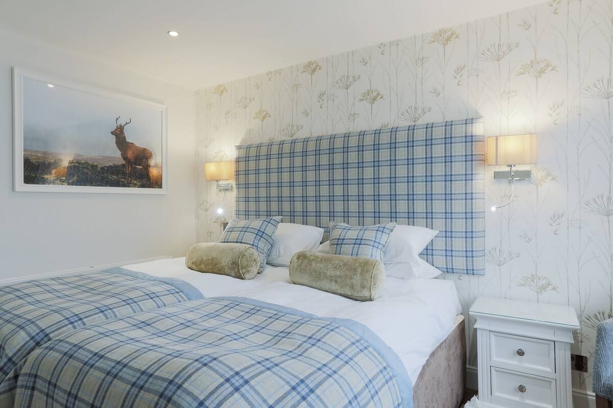 A bedroom with a large bed, plaid bedding, a mounted deer picture, and a nightstand with a lamp.
