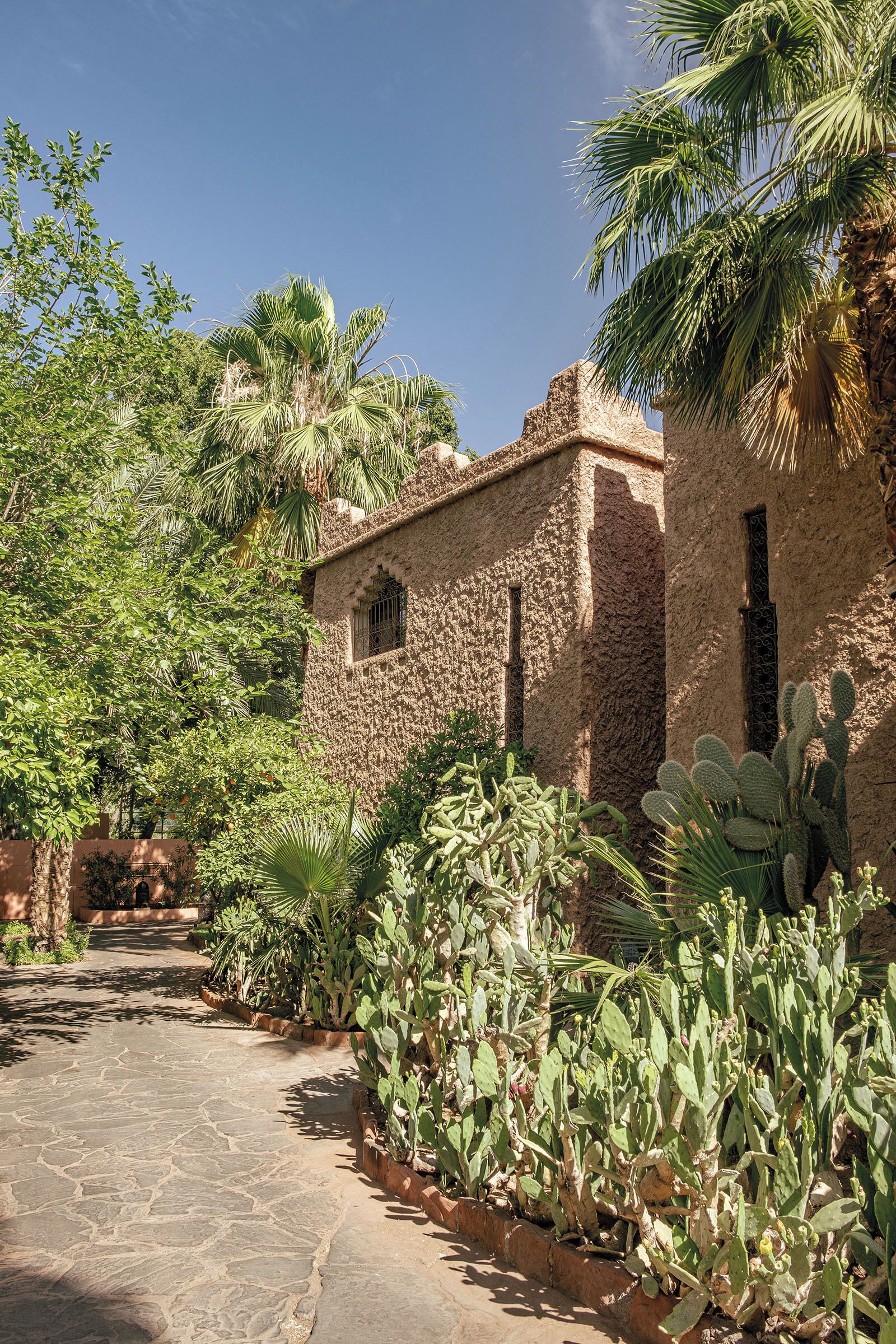 Un bâtiment en pierre, avec un étroit sentier également en pierre, entouré de cactus et de palmiers.