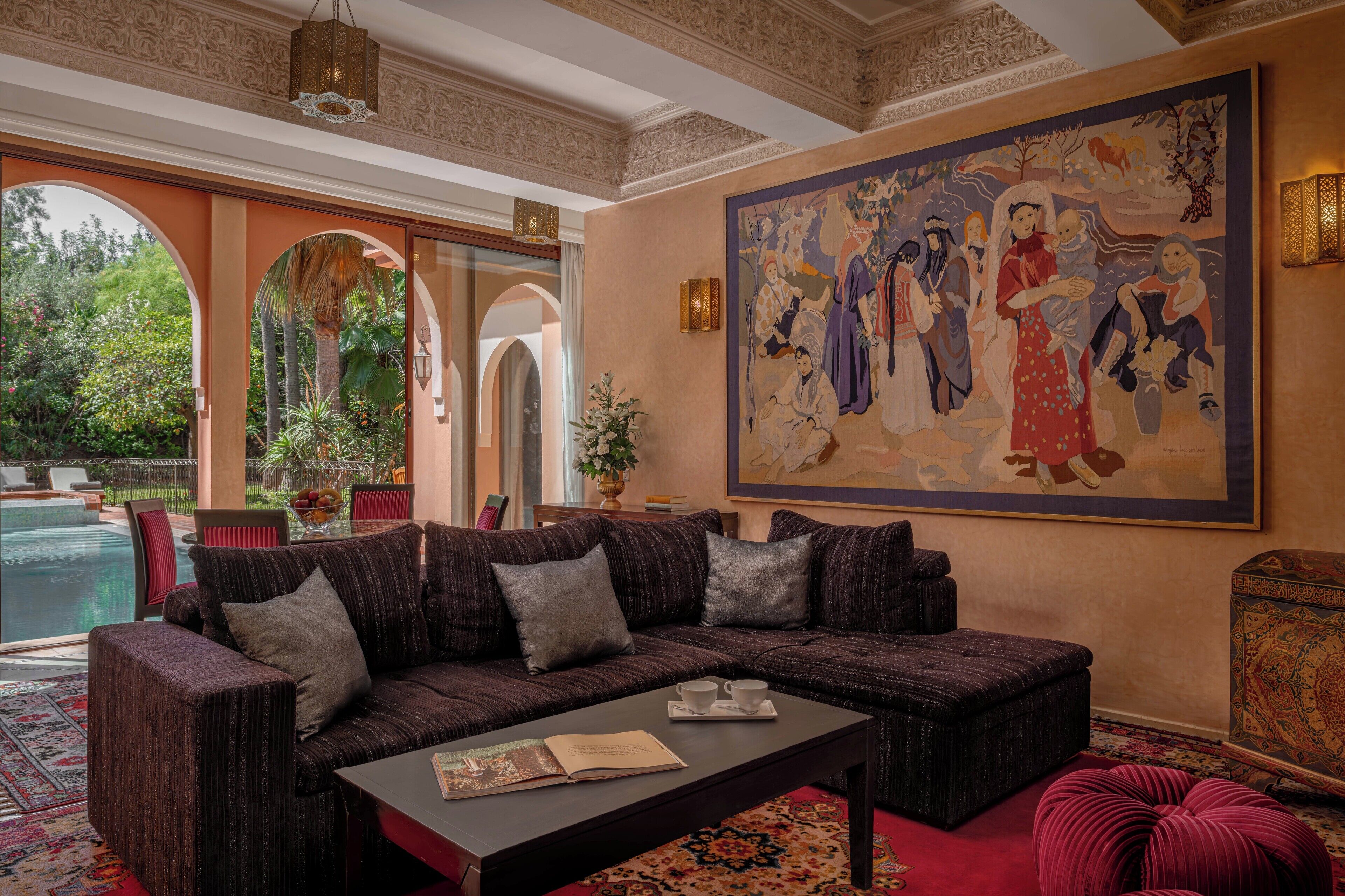 A living room with a sofa, a coffee table, and a large painting. There is a pool visible through an archway.