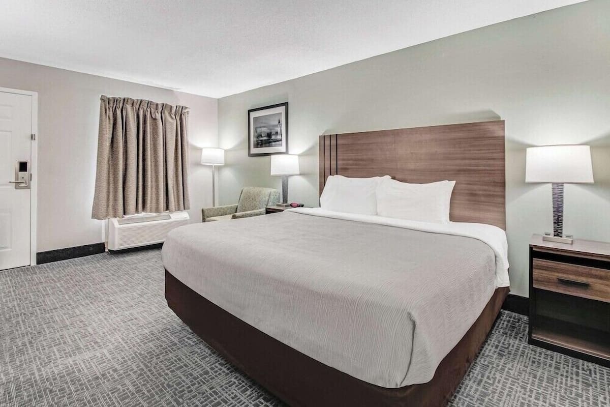 Habitación de hotel con una cama grande, dos mesitas de noche con lámparas, una silla, una ventana con cortinas y una puerta.