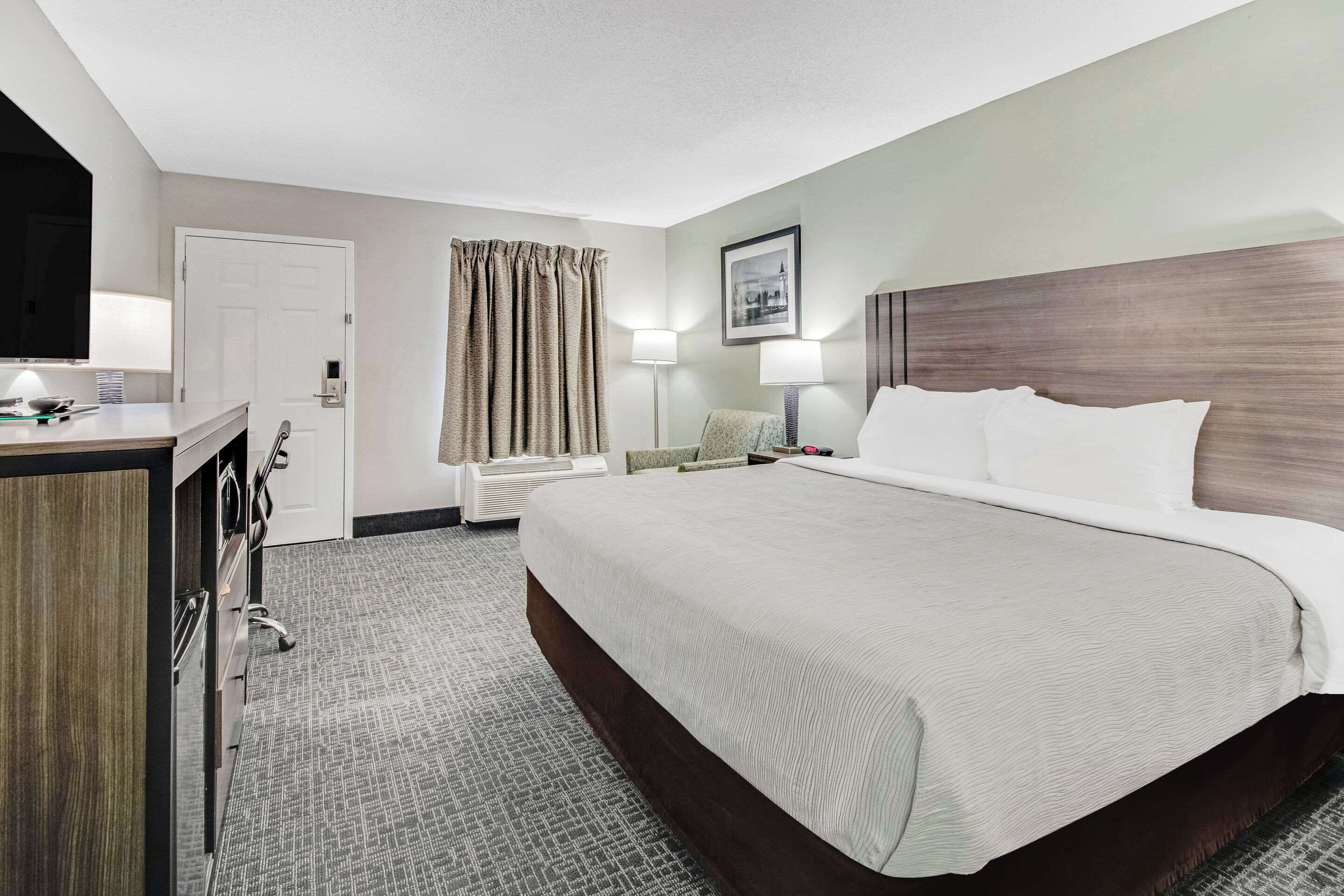A hotel room with a large bed, a desk with a lamp, a chair, a wardrobe, and a window with curtains.