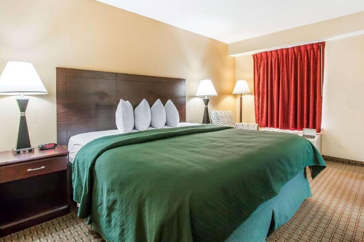 Habitación de hotel con cama, dos lámparas de noche, una mesita de noche, una cortina roja y un reloj digital.